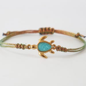 Może przedstawiać: Bransoletka ze złotym urokiem żółwia i turkusowym środkiem. Bransoletka posiada pleciony sznurek w odcieniach zieleni i brązu. Urok żółwia ma około 2,5 cm długości.