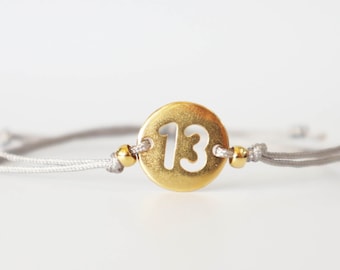 May include: A gray string bracelet with a gold circle charm featuring the number 13 in white.