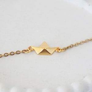 Könnte beinhalten: Halskette mit einem goldfarbenen Papierboot-Anhänger. Der Anhänger ist ein kleines Boot im Origami-Stil, das an einer zarten Kette hängt. Die Halskette wird auf einer weißen Oberfläche mit gewelltem Rand präsentiert.