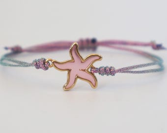 Puede incluir: Una pulsera con un dije central de estrella de mar en esmalte rosa con bordes dorados. El dije está sujeto a un cordón fino y ajustable hecho de hilos tejidos en tonos morado y azul, anudados a ambos lados de la estrella de mar.