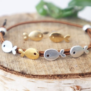 May include: A brown leather bracelet with silver and gold fish charms. The bracelet is on a wooden surface.