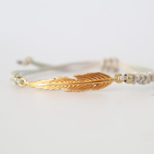 May include: A gold feather charm bracelet with a braided cord in grey and white. The feather is the focal point, with detailed veining. The bracelet is adjustable.
