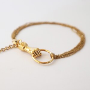 Pode incluir: Pulseira dourada com um fecho detalhado em forma de punho segurando um anel. A pulseira tem várias correntes delicadas e um fecho de corrente ajustável. A joia está sobre um fundo branco liso, destacando o design intrincado e a cor quente.
