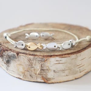 Puede incluir: Un brazalete con un cordón beige y abalorios de peces plateados y dorados. El brazalete está sobre una superficie de madera.