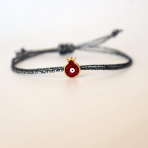 Könnte beinhalten: Ein zartes Armband mit einem roten Granatapfel-Anhänger mit goldfarbener Umrandung und einem kleinen weißen Augendesign. Der Anhänger ist an einem dünnen, dunkelgrauen Band befestigt. Das Armband ist verstellbar.