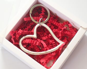 May include: A silver-toned metal heart-shaped keychain with a circular split ring attachment, nestled inside a white square gift box filled with shredded red paper filler. The heart outline is smooth and simple, providing a clean aesthetic.
