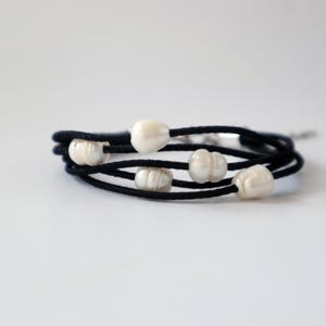 Peut inclure: Bracelet à plusieurs brins avec cinq perles blanches de forme irrégulière sur un cordon noir. Le bracelet est enroulé, créant un effet de superposition. Les perles ont un éclat brillant et le cordon est en matière douce et mate. Bijou.