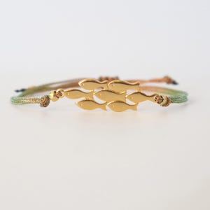 Puede incluir: Pulsera dorada con diseño de peces. La pulsera tiene un cordón tejido en tonos verdes, marrones y dorados. Los peces están dispuestos en un patrón de natación. Un accesorio de temática náutica.