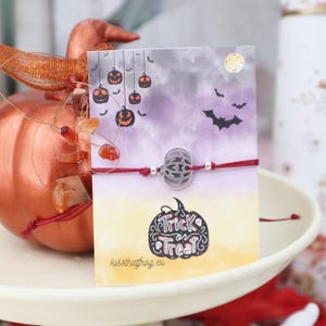 Op de afbeelding: Een rood koordarmbandje met een zilverkleurige pompoenbedel, gepresenteerd op een rechthoekige kaart met een paars en geel verloop, illustraties van hangende pompoenen, vleermuizen en de tekst Trick or Treat en kissthatfrog.eu.