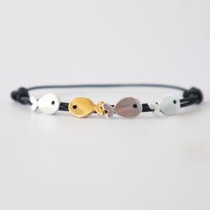 Könnte beinhalten: Schwarzes Kordelarmband mit vier kleinen Fischanhängern. Die Anhänger sind in Silber- und Goldtönen gehalten und haben ein minimalistisches Design. Das Armband hat einen Knotenverschluss.