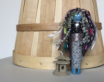 May include: A blue and white doll with long, colorful hair and a silver heart necklace. The doll is wearing a silver and black polka dot skirt and a silver belt. The doll is standing next to a small, gray, pagoda-style building.