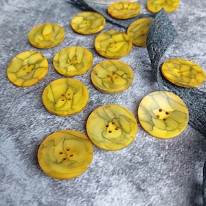 Puede incluir: Una colección de 12 botones amarillos y verdes con un patrón de mármol. Los botones tienen cuatro agujeros cada uno y están dispuestos en un semicírculo sobre una superficie gris.