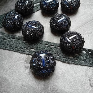 Puede incluir: Primer plano de ocho botones decorativos azul oscuro con detalles intrincados. Cada botón tiene forma redonda con superficie texturizada y está adornado con pequeños acentos brillantes. Una cinta de encaje gris oscuro y una bobina de madera en el fondo.