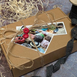 Peut inclure: Une boîte en carton marron attachée avec de la ficelle, ornée d'une petite photo et de boutons rouges. La boîte est décorée d'une photo d'objets colorés. Une bobine de dentelle noire et du matériau en forme de paille sont également présents.