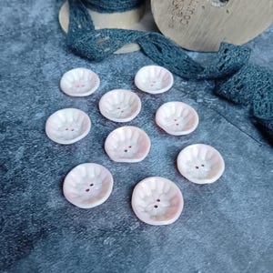 Pode incluir: Uma coleção de dez botões cor-de-rosa com um design em relevo festoneado e dois furos cada. Os botões estão dispostos em semicírculo sobre uma superfície cinzenta.