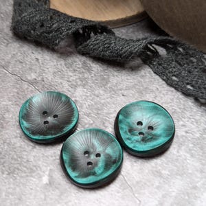 Puede incluir: Tres botones verdes y negros con cuatro agujeros cada uno. Los botones tienen un acabado brillante e iridiscente.