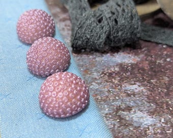 May include: Three dome-shaped buttons in a dusty mauve color, each featuring a raised, circular, honeycomb-like texture across the surface. The buttons are arranged in a vertical line, resting on a light blue fabric with a subtle cross-stitch pattern.
