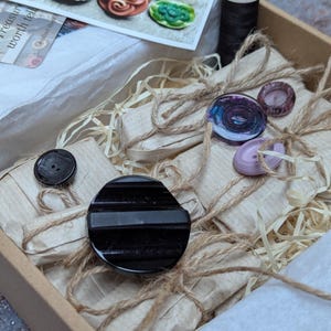 May include: A craft box containing an assortment of colourful buttons, wrapped parcels, and a spool of black thread. The buttons are in shades of orange, green, blue, and pink. The parcels are tied with twine.