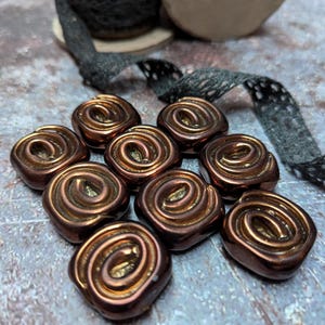 May include: Eight square-shaped buttons with a metallic bronze finish and a raised spiral design on the surface. The buttons are arranged in a cluster, accompanied by a spool of black lace trim with a delicate, intricate openwork pattern.