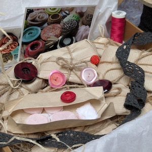 Könnte beinhalten: Eine Kiste voller Nähzubehör. Sie enthält verpackte Päckchen, Knöpfe in Rosa, Rot und Braun, eine Spule rosa Garn und graue Spitzenborte. Ein Foto von Knöpfen ist ebenfalls zu sehen.