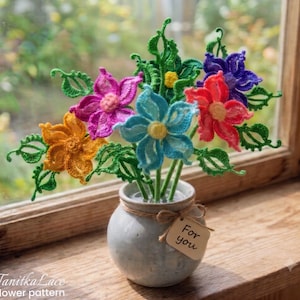 May include: A bouquet of crocheted flowers in orange, pink, blue, red, and purple with green leaves and stems, arranged in a small, round, light gray ceramic vase. A small, light brown rectangular tag tied with twine reads "For you".