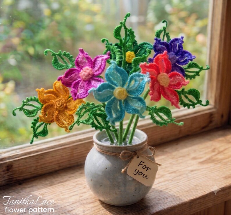 May include: A bouquet of crocheted flowers in orange, pink, blue, red, and purple with green leaves and stems, arranged in a small, round, light gray ceramic vase. A small, light brown rectangular tag tied with twine reads "For you".