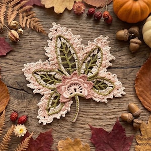 May include: A handmade crochet leaf in shades of pink, green, and cream, with intricate openwork details. The leaf is surrounded by autumn leaves, acorns, and small pumpkins on a rustic wooden surface. The crochet pattern is visible.