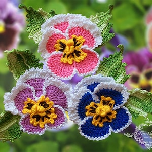 May include: Three crocheted pansy flowers with green leaves. The flowers are pink, purple, and blue with white edges. The flowers are on a green background.