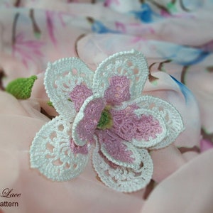 May include: A handmade crochet flower with white, pink, and green accents. The flower has a detailed openwork design and is set against a soft, patterned fabric background. The text "Tanitka Lace flower pattern" is visible.
