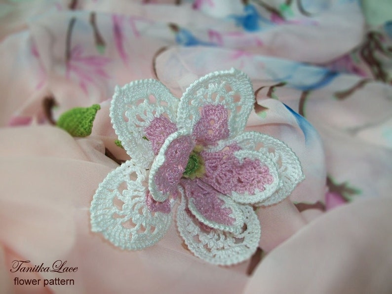 May include: A handmade crochet flower with white, pink, and green accents. The flower has a detailed openwork design and is set against a soft, patterned fabric background. The text "Tanitka Lace flower pattern" is visible.