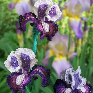 May include: Handmade crochet iris flowers in shades of purple, white, and green. The flowers have detailed petal work and are displayed against a blurred background of similar flowers. The text "Tanitka Lace flower pattern" is visible.