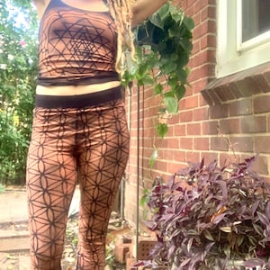 May include: A woman wearing a brown and black tank top and leggings with a geometric pattern. The leggings are tight-fitting and have a high waist. The tank top is sleeveless and has a scoop neck. The woman is standing in front of a brick wall with plants in the background.