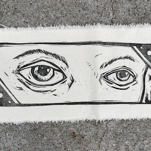 Könnte beinhalten: Schwarz-weißer Linolschnitt von zwei Augen auf weißem Stoffhintergrund. Die Augen sind in einem schwarzen Rechteck mit geometrischem Muster gerahmt.