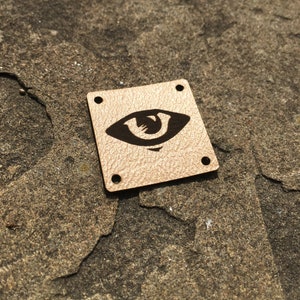 May include: A square-shaped leather patch with a brown eye design. The patch has four holes around the edges.