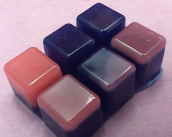 May include: Six square-shaped blocks of soap, each with a different color combination. The blocks are arranged in a staggered pattern, with the top block being purple and the bottom block being pink.