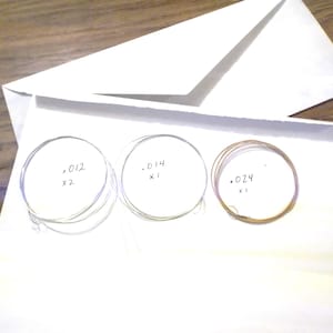 May include: Three sets of guitar strings on a white surface. The strings are labeled with their gauge and quantity: .012 x2, .014 x1, and .024 x1.
