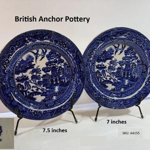 May include: Two blue and white British Anchor Pottery plates, one 7.5 inches and the other 7 inches in diameter, displayed on black stands. The plates feature a detailed landscape design. The text "British Anchor Pottery" is visible.