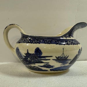 May include: Cream-colored ceramic pitcher with a blue Willow pattern. The pitcher features a curved handle and a spout. The design includes a pagoda, boat, and landscape scenes. The rim and handle are decorated with floral patterns.