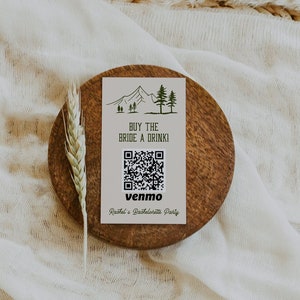 May include: A white card with a mountain and trees illustration, the text "Buy the Bride a Drink!" and a QR code with the text "Venmo" and "Ruth's Bachelorette Party". The card is on a brown wooden coaster.