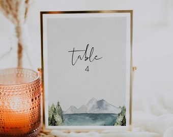 Op de afbeelding: Een gouden gelijst tafelnummerkaart met een aquarel-illustratie van een bergketen en een meer. De kaart heeft de tekst "tafel 4" en ligt op een geweven placemat met een kaars op de voorgrond.