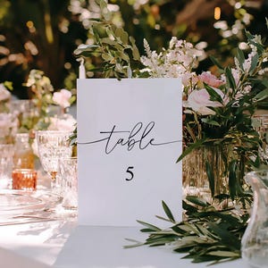 May include: A white table card with the word "table" in cursive and the number "5" in a black font. The card is on a white tablecloth, surrounded by floral arrangements, glassware, and candles, creating an elegant setting.