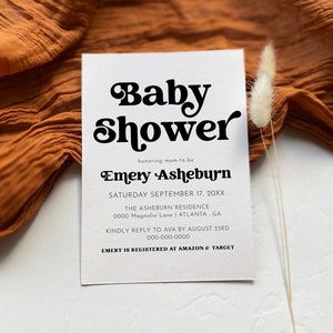 Op de afbeelding: Een witte babyborrel-uitnodiging met zwarte tekst. De uitnodiging luidt "Baby Shower" in een retro-lettertype, gevolgd door "honoring mom-to-be Emery Asheburn". De datum, tijd en locatie van de borrel staan hieronder vermeld. De uitnodiging bevat ook een telefoonnummer en een notitie dat Emery is geregistreerd bij Amazon en Target.