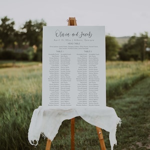 May include: A white wedding seating chart with the names Olivia and Jacob at the top. The chart lists the names of guests assigned to each table. The chart is displayed on a wooden easel with a white cloth draped over it. The chart is set up outdoors in a grassy field.