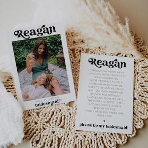 May include: A white card with black text that reads "Reagan" in a retro font. The card has a photo of two women smiling and holding glasses of wine. The text below the photo reads "Will you be my bridesmaid?"