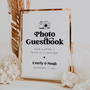 May include: A gold-framed sign with a white background and black text that reads "Photo Guestbook" with a camera icon. The text below reads "snap a photo + leave us a message!" and "Everly & Noah" with the date "NOVEMBER 11, 20xx".