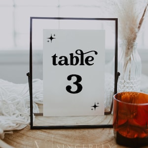 May include: Black and white table number sign with the text "table 3" in a black frame. The sign is sitting on a wooden surface with a white fuzzy blanket and a glass vase with dried pampas grass.