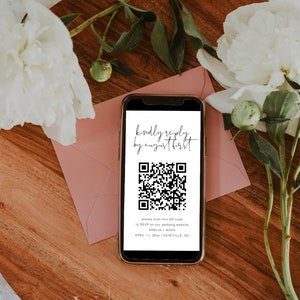 May include: A smartphone displaying a QR code with the text "kindly reply, by august first" and instructions to RSVP on a wedding website for Amelia + Noah on April 11, 20xx in Asheville, NC. The phone is on a wooden surface with a pink envelope and white flowers.