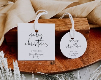 Könnte beinhalten: Zwei weiße Geschenkanhänger mit schwarzer Kalligraphie, auf denen "Frohe Weihnachten" und "Mit Liebe, die Familie Williamson" steht. Die Anhänger befinden sich auf einer Holzoberfläche mit einem braunen Band.