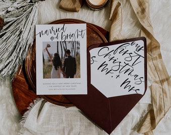 May include: A white Christmas card with a photo of a couple in a forest. The card reads "married and bright" and "Wishing you a very Merry Christmas - The Happiest of New Years Love Brit & Oliver Williamson". The card is on a wooden plate with a brown ribbon and a candle.