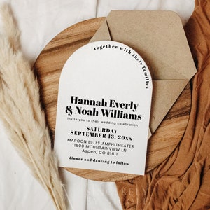 May include: A white wedding invitation with a rounded arch shape. The invitation reads "Hannah Everly & Noah Williams" and includes the wedding date, location, and time. The invitation is on a wooden surface with a brown envelope and a bouquet of dried pampas grass.